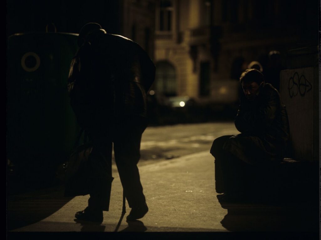 Karol (Zbigniew Zamachowski) sits on the sidewalk. Next to him, a man throwing a bottle (played by the director Kieślowski).