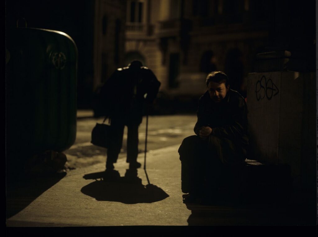 Karol (Zbigniew Zamachowski) sits on the sidewalk. Next to him, a man throwing a bottle (played by the director Kieślowski).