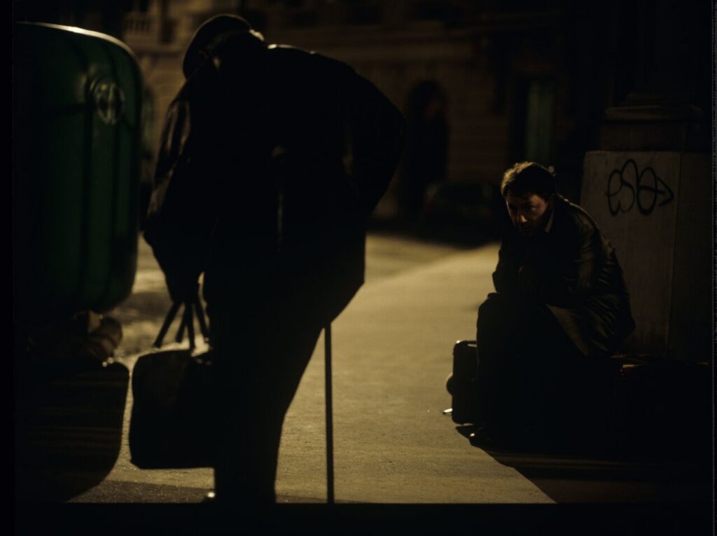 Karol (Zbigniew Zamachowski) sits on the sidewalk. Next to him, a man throwing a bottle (played by the director Kieślowski).