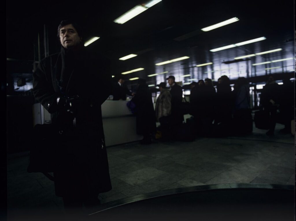 Mikołaj (Janusz Gajos) is waiting for his luggage in the arrivals hall at the Warsaw airport.