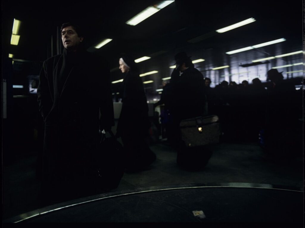 Mikołaj (Janusz Gajos) is waiting for his luggage in the arrivals hall at the Warsaw airport.