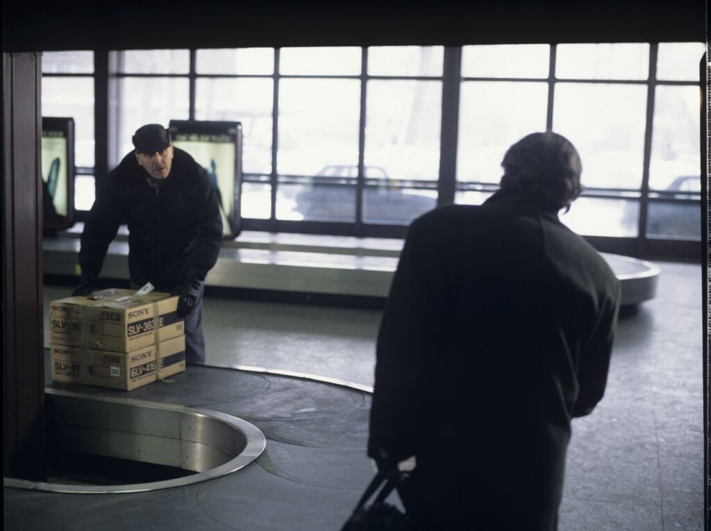 Mikołaj (Janusz Gajos) is waiting for his luggage in the arrivals hall at the Warsaw airport.