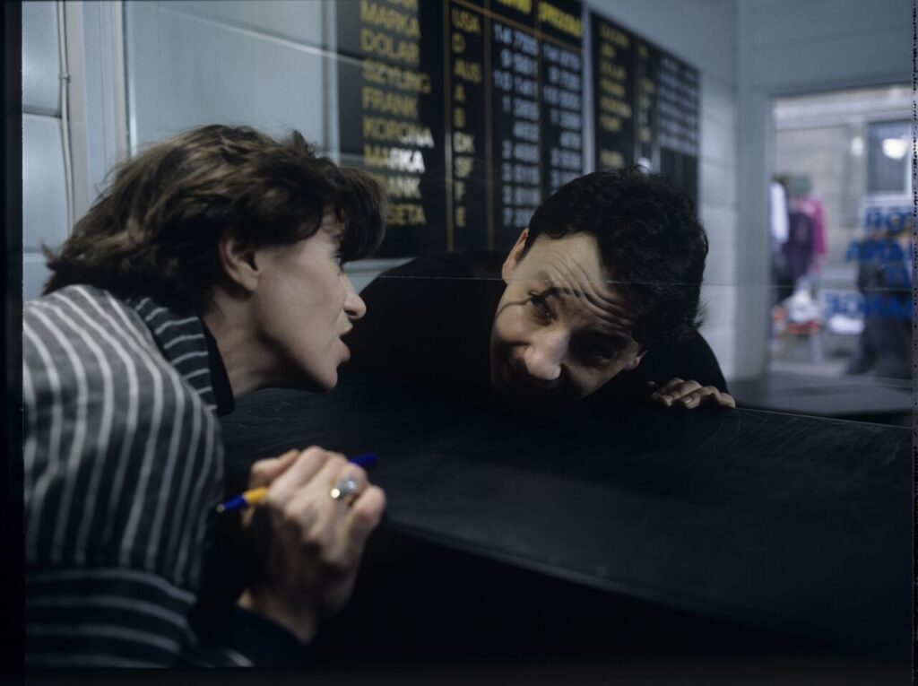 Karol (Zamachowski) talks to the cashier (Dean) of the currency exchange office. The scene was not shown in the film.