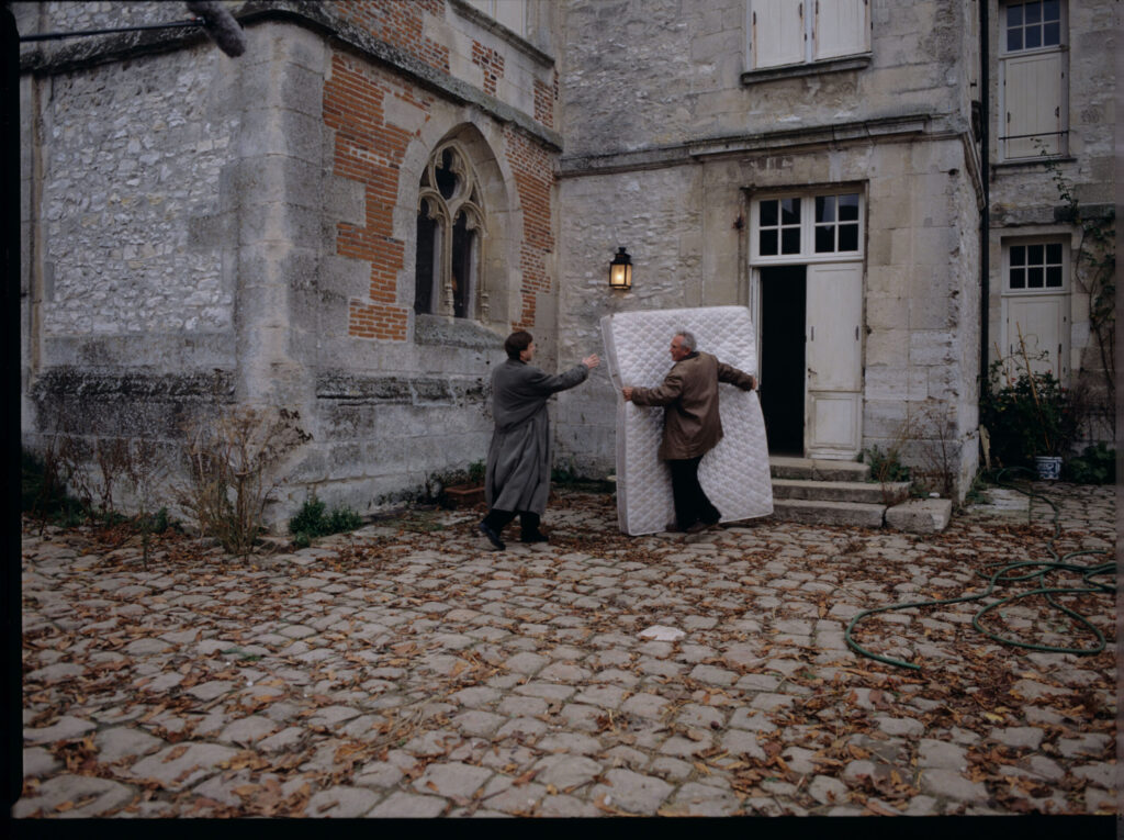 Oliviere (Régent) carries the mattress to the car with the help of a gardener (Forget). The scene was not shown in the film.