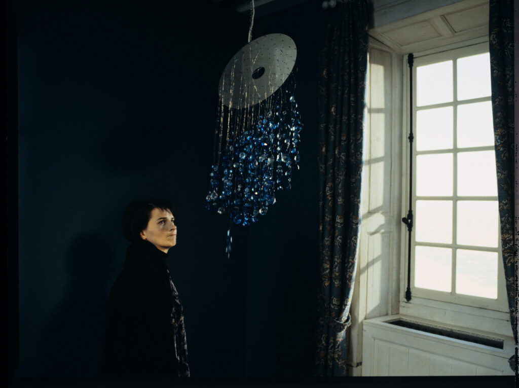 Julie (Juliette Binoche), in the room of her deceased daughter, angrily pulls the glass of the blue chandelier left behind.