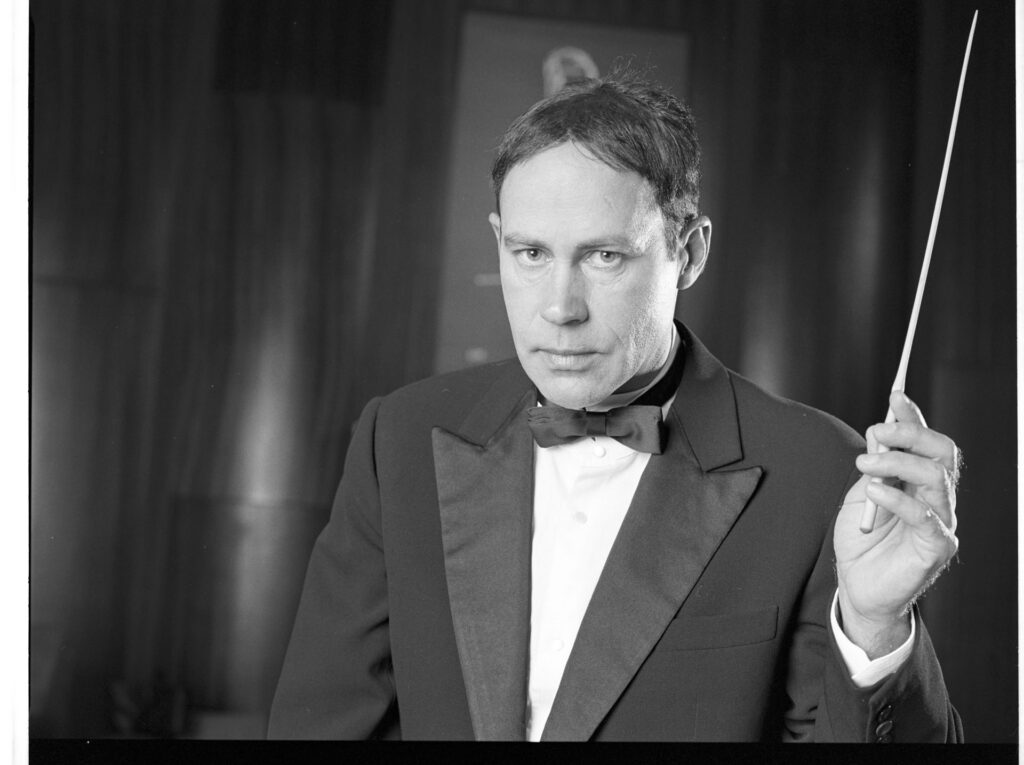 A photograph of the composer Patrice de Courcy (Hugues Quester) rehearsing with the orchestra.