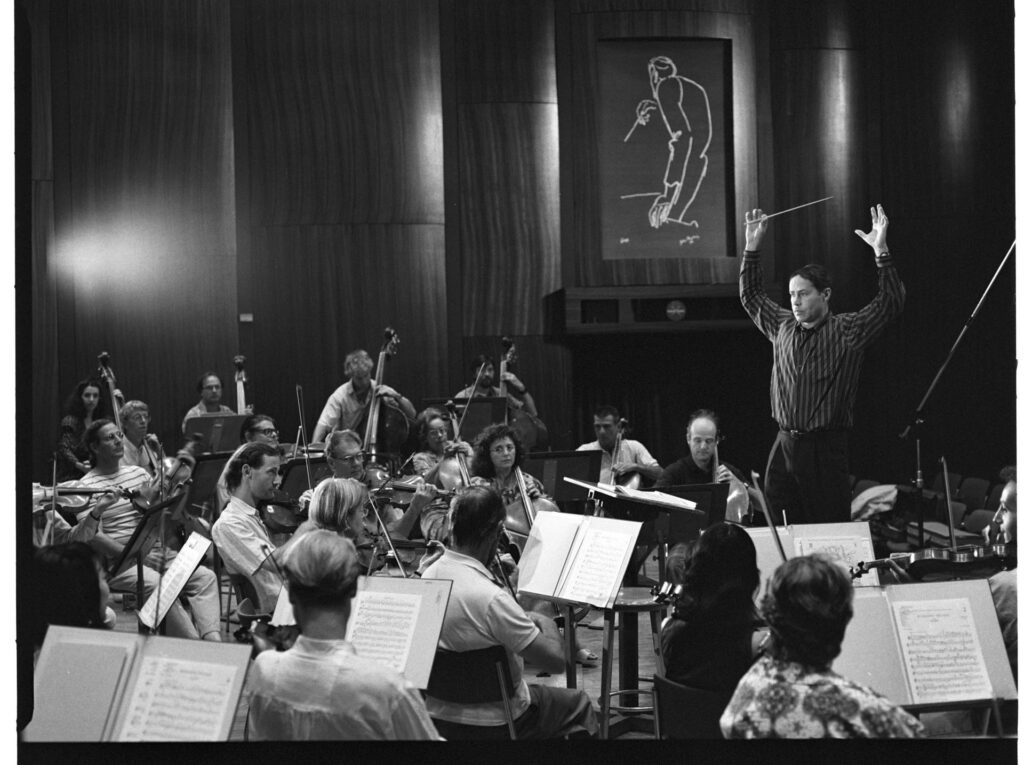 A photograph of the composer Patrice de Courcy (Hugues Quester) rehearsing with the orchestra.