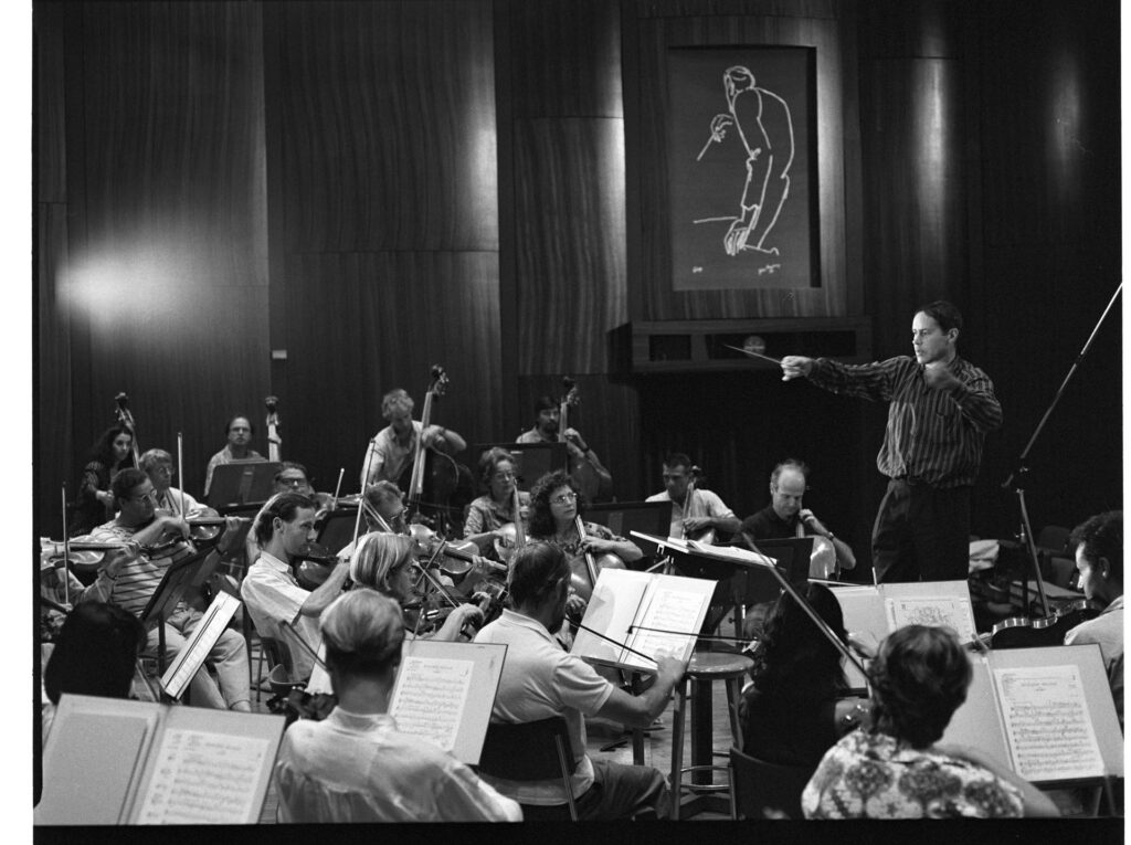 A photograph of the composer Patrice de Courcy (Hugues Quester) rehearsing with the orchestra.