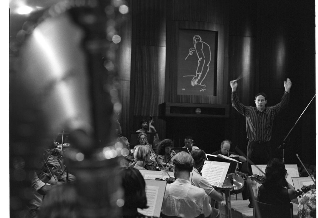A photograph of the composer Patrice de Courcy (Hugues Quester) rehearsing with the orchestra.